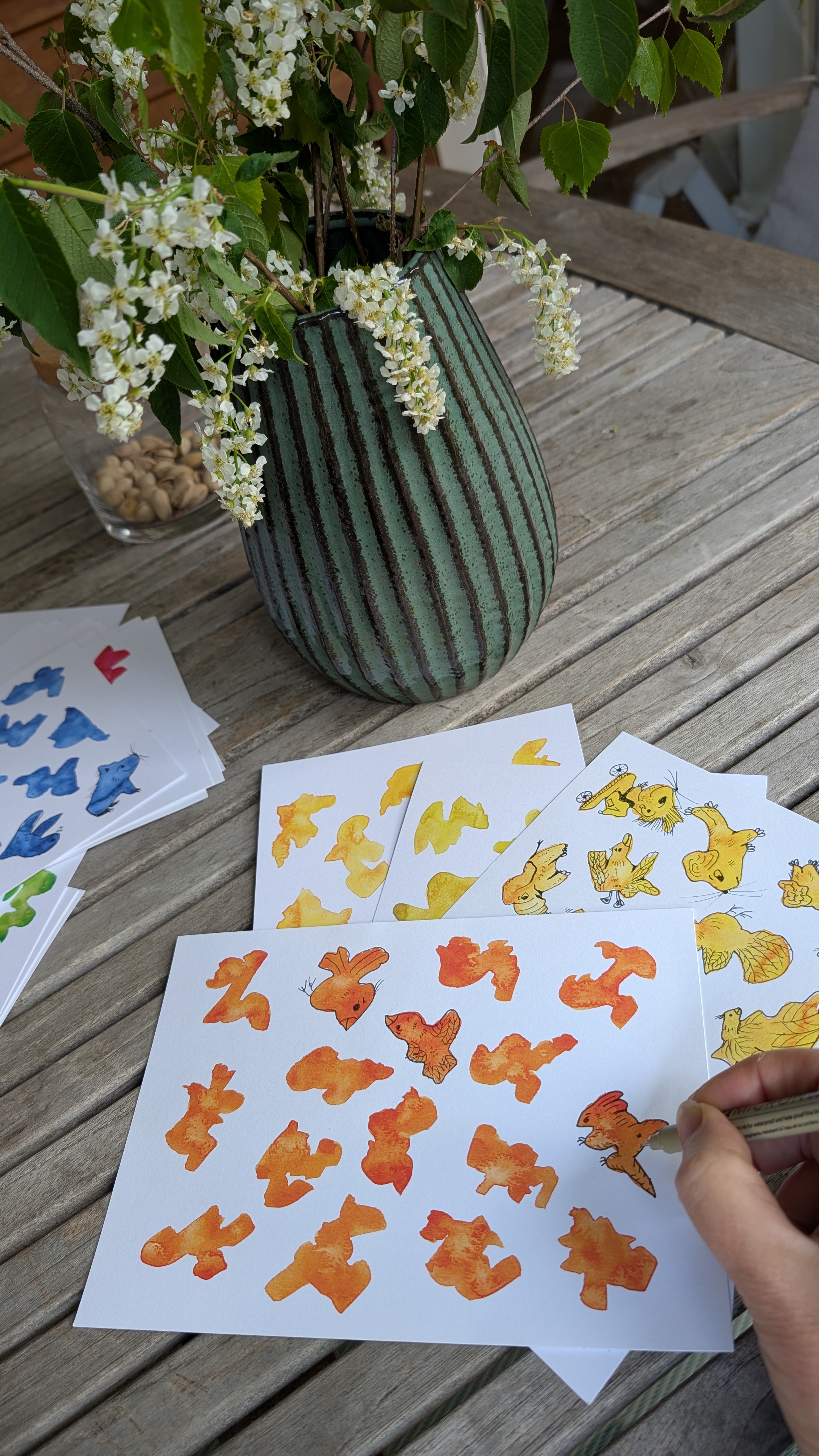 A hand doodling on an orange watercolor card at an outdoor table, with several colorful cards nearby.